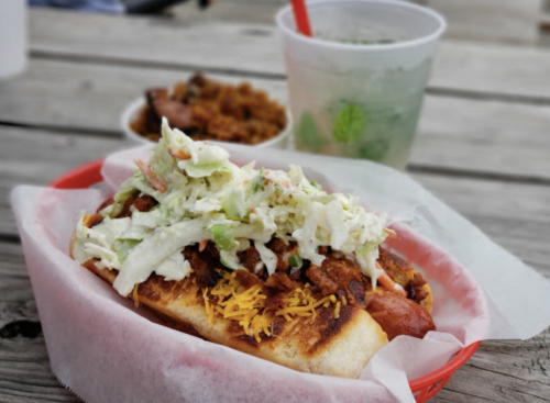 A hot dog topped with chili, cheese, and coleslaw, served in a basket with a drink and side dish in the background.