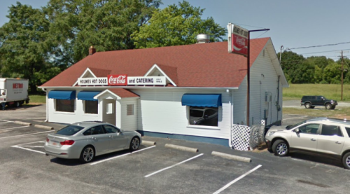 A white building with a red roof, featuring blue awnings and a sign for "Holmes Hot Dogs and Catering," surrounded by parked cars.