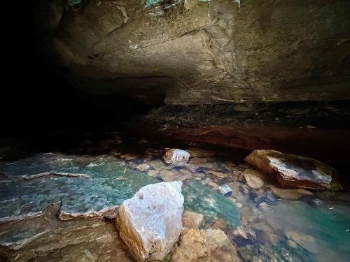 A dark cave interior with a shallow pool of water and large rocks scattered on the floor.