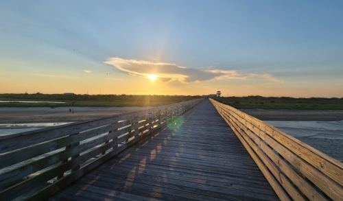A wooden pier stretches into the distance under a sunset, with soft clouds and a serene landscape.