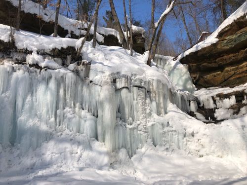 Icicles and frozen waterfalls cascade down a snowy cliff, surrounded by trees under a clear blue sky.