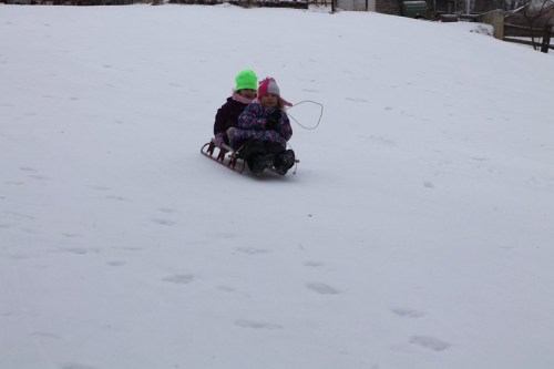 Two children in winter clothing sledding down a snowy hill, smiling and enjoying the winter day.