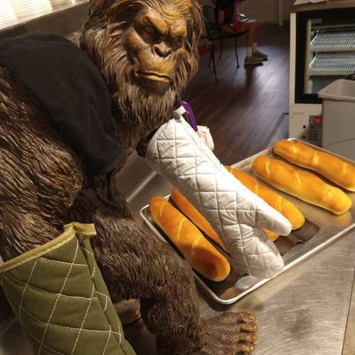 A large, furry creature in an oven mitt pulls fresh bread from a tray in a kitchen setting.