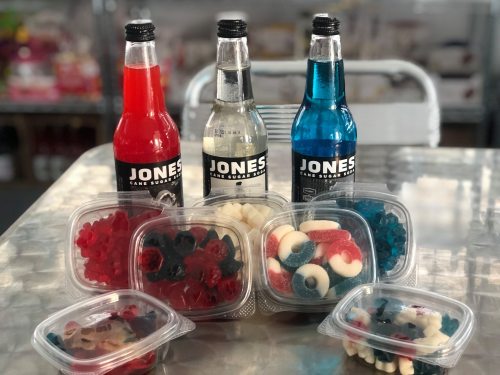 Three colorful Jones soda bottles with red, white, and blue gummy candies in clear containers on a metallic surface.