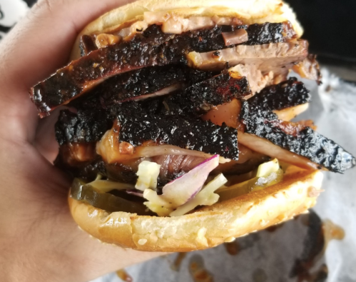 A hand holding a stacked sandwich filled with layers of smoked meat, pickles, and coleslaw on a sesame seed bun.