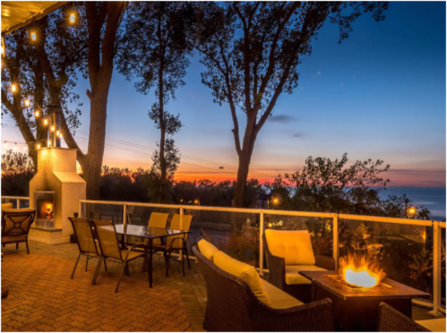 Cozy outdoor seating area with a fire pit, overlooking a sunset by the ocean, surrounded by trees and string lights.