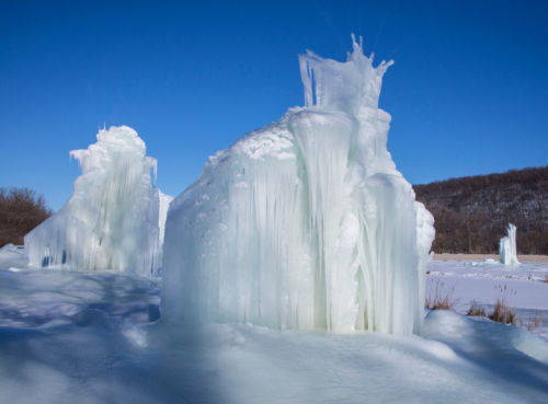 Ice formations rise majestically against a clear blue sky, surrounded by snow-covered ground and distant hills.