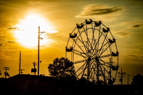 Silhouette of a ferris wheel against a vibrant sunset sky, with power lines and trees in the foreground.