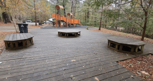 Wooden deck with two circular benches, surrounded by trees, with an orange playground structure in the background.