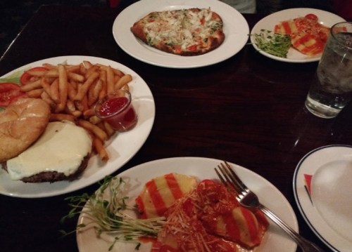 A table with a burger and fries, a pizza, and a plate of pasta with sauce and cheese, alongside a glass of water.