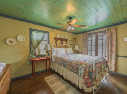 Cozy bedroom with a floral bedspread, wooden floor, green ceiling, and vintage decor, featuring a window and lamps.