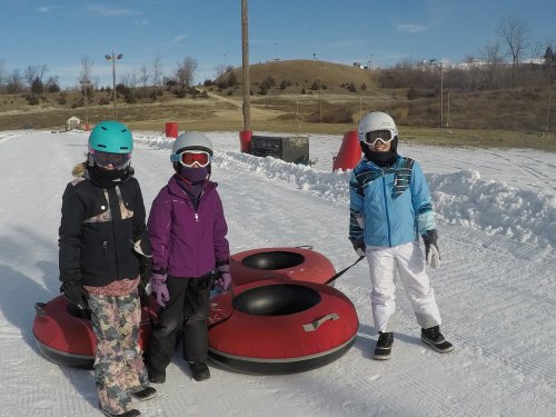 Three children in winter gear stand beside red snow tubes on a snowy slope, with a hill and blue sky in the background.