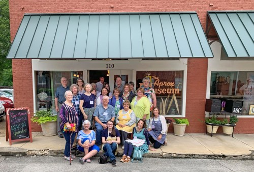 A group of people gathered outside The Apron Museum, smiling and posing for a photo in front of the building.