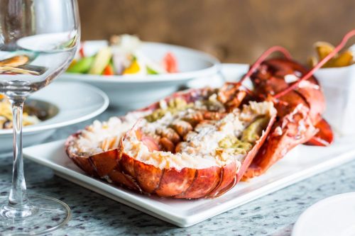 A beautifully presented lobster on a white plate, accompanied by a glass of wine and side dishes in the background.
