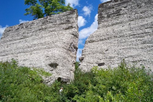 Two tall, weathered rock formations split by a narrow gap, surrounded by lush greenery and a bright blue sky.