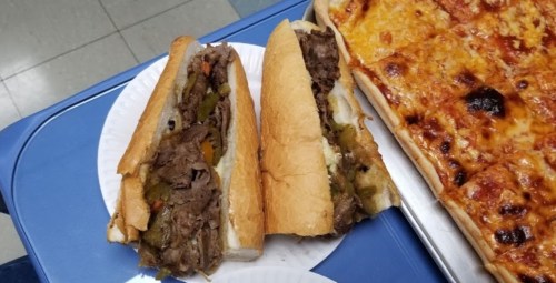 Two large sandwiches filled with beef, peppers, and onions, alongside a tray of cheesy pizza.