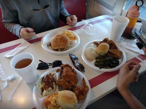 Two plates of Southern food with fried chicken, burgers, sides, and drinks on a table. Sunglasses and utensils are nearby.