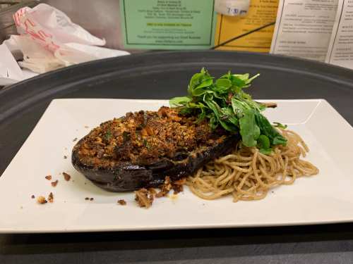 Stuffed eggplant on a plate, topped with greens, served alongside a portion of noodles.