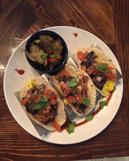 Three tacos filled with meat, pico de gallo, and cilantro, served with a side of seasoned potatoes on a wooden table.