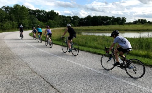 A group of cyclists riding along a paved path beside a grassy area and a pond under a partly cloudy sky.