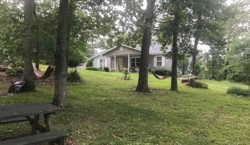 A cozy house surrounded by trees, with two hammocks and a picnic table in a lush green yard.