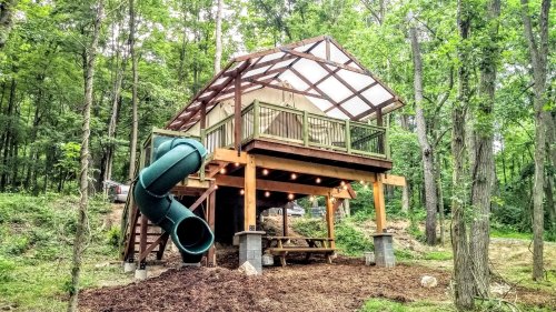 A treehouse with a slide, elevated on stilts, surrounded by trees and featuring a deck and picnic table below.