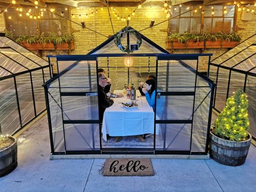A cozy outdoor dining setup in a greenhouse with two people at a table, surrounded by lights and greenery.