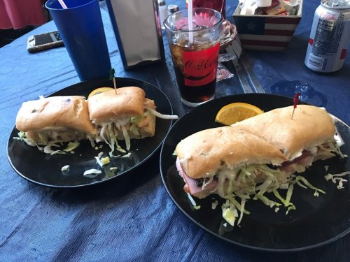 Two sandwiches on black plates, filled with lettuce and ham, accompanied by a glass of cola and a slice of orange.