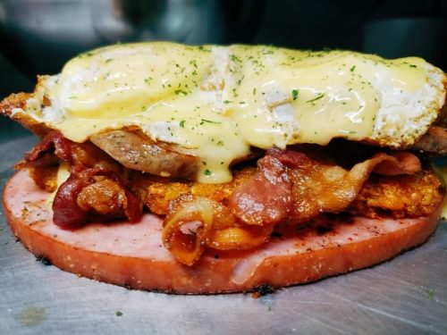 A hearty sandwich with layers of bacon, sausage, fried egg, and creamy sauce on a slice of ham.