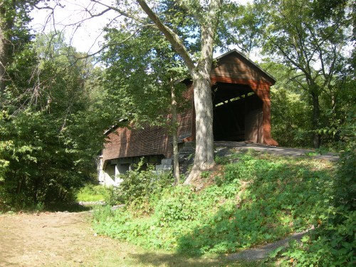 A wooden covered bridge surrounded by trees and greenery, set on a gentle slope.