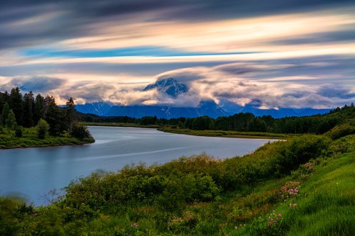 A serene river flows through lush greenery, with mountains partially shrouded in clouds under a colorful sky.