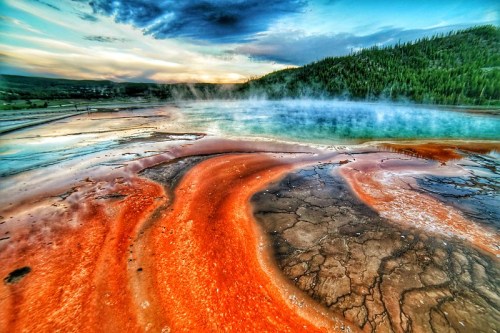 Vibrant geothermal pool with orange and blue hues, surrounded by steam and lush greenery under a colorful sky.