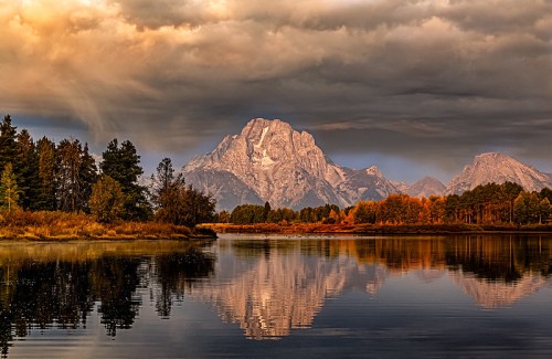 A serene lake reflects a majestic mountain under a dramatic sky, surrounded by trees and autumn foliage.