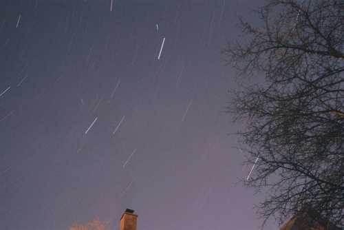 Star trails streak across a night sky, with a silhouette of a tree and a chimney in the foreground.