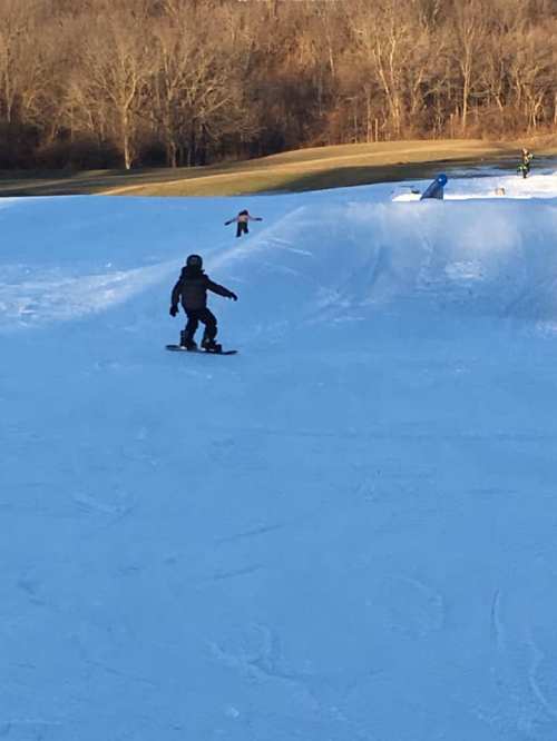 A child snowboards on a snowy slope, while another child plays in the background. Trees line the area.