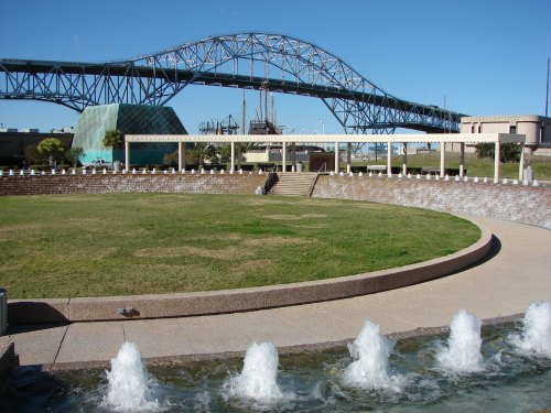 A park with a circular fountain and grassy area, featuring a large blue bridge in the background under a clear sky.