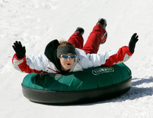 A person in winter gear slides down a snowy hill on a green snow tube, arms raised in excitement.