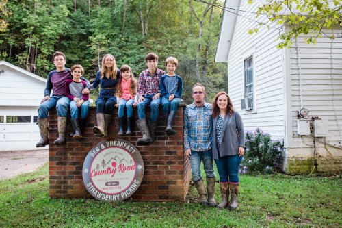 A family poses on a brick wall sign for "Country Road B&B" with children sitting above and adults standing below.