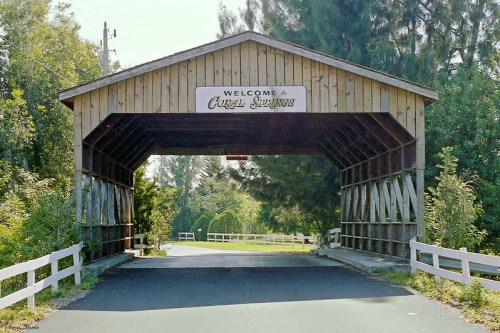 A wooden covered bridge with a sign reading "Welcome to Coral Springs," surrounded by greenery and a paved road.