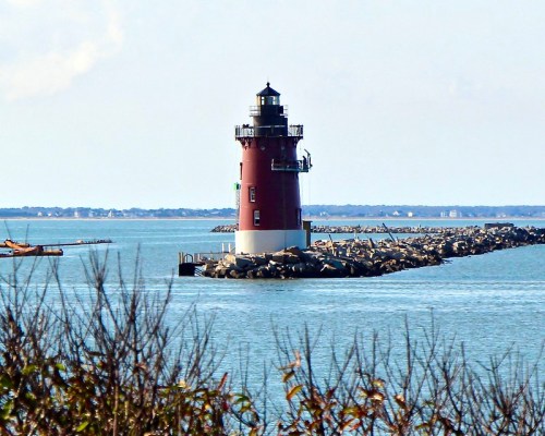 A red and white lighthouse stands on a rocky jetty, surrounded by calm blue waters and a clear sky.
