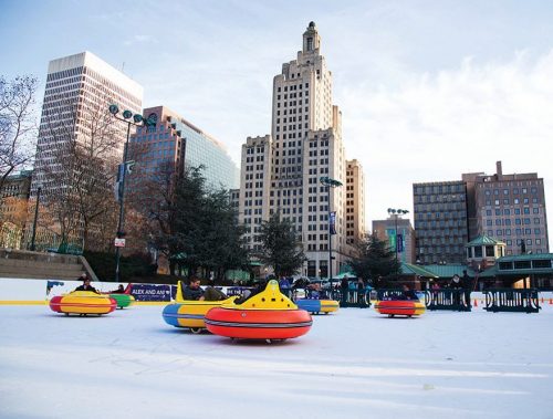 Colorful bumper cars on an ice rink with city buildings in the background under a clear sky.