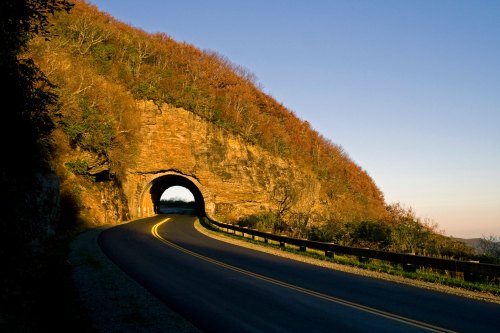 A winding road leads through a rocky tunnel, surrounded by autumn foliage and a clear blue sky.