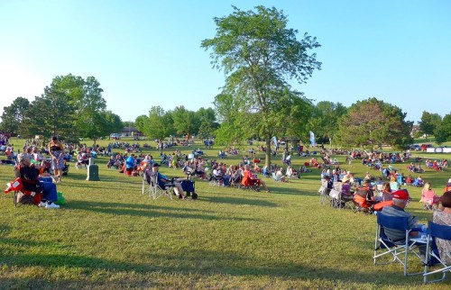 A large crowd of people sitting on lawn chairs in a park, enjoying a sunny day with trees in the background.