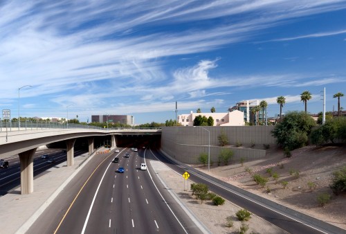 A view of a highway with vehicles, under a blue sky with clouds, and buildings and palm trees in the background.