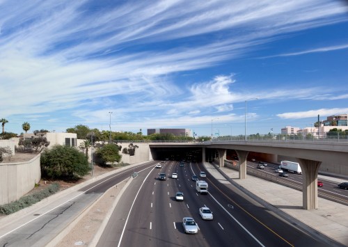 A view of a busy highway with cars, under a clear blue sky with wispy clouds, and a concrete overpass above.