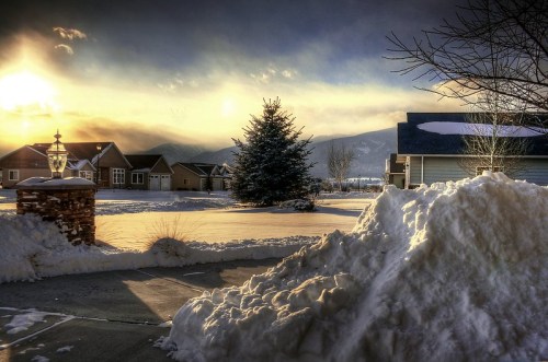 A snowy landscape at sunset, featuring houses, a snow-covered driveway, and a tall evergreen tree.