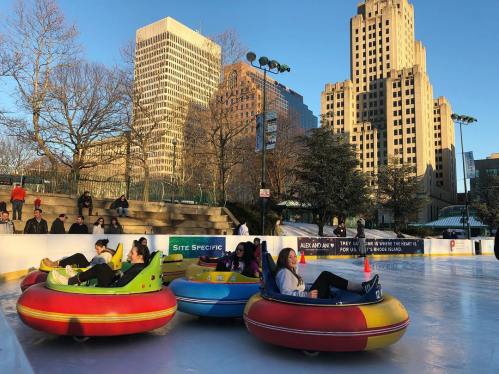 People enjoying a winter ice skating rink in colorful inflatable tubes, with city buildings in the background.