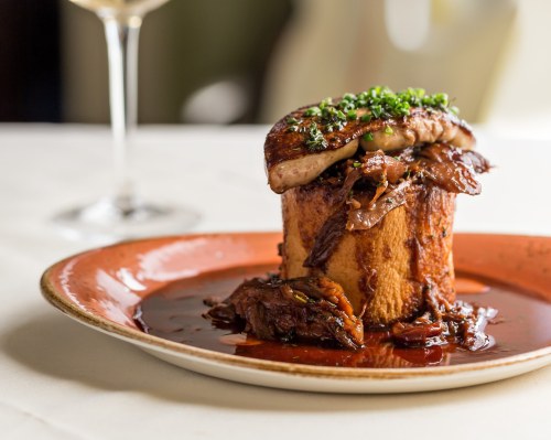 A gourmet dish featuring a savory meat stack on a bread base, garnished with herbs and served in a rich sauce.