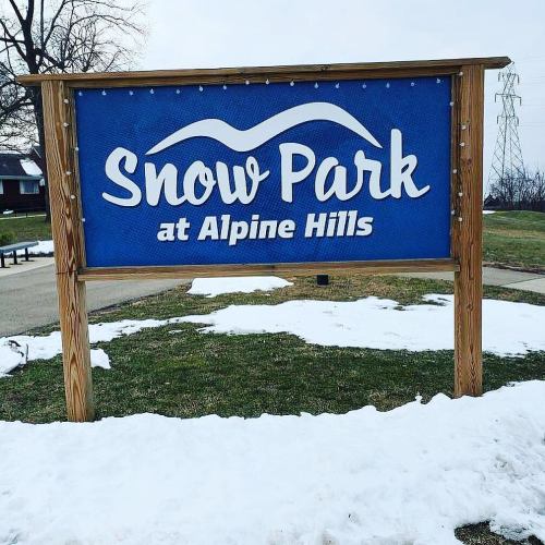 Sign for Snow Park at Alpine Hills, featuring a blue background and white text, with snow on the ground nearby.
