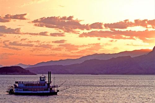 A steamboat sails on a calm lake at sunset, with mountains silhouetted against a colorful sky.